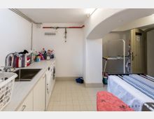 Waschraum mit gefliesten Böden und weiß gestrichenen Wänden. Links befinden sich eine Arbeitsplatte mit Spüle, Waschmaschine, Trockner und Reinigungsmitteln, während rechts ein Wäscheständer und eine Tür sichtbar sind.