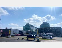 Ein moderner Gewerbekomplex mit Glasfassaden im Hintergrund, davor ein Parkplatz mit mehreren Autos sowie Stapel von Baumaterialien und ein Flachbau mit Werbeschildern. Der Himmel ist teils bewölkt mit sichtbarem Sonnenlicht.