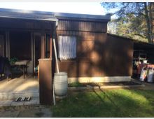 Holzlaube mit dunkelbrauner Holzfassade und einem kleinen, überdachten Eingangsbereich. Vor der Laube befindet sich eine Grünfläche, daneben steht eine Regentonne und ein Blumenbeet ist angelegt. Im Hintergrund sind Bäume zu sehen.