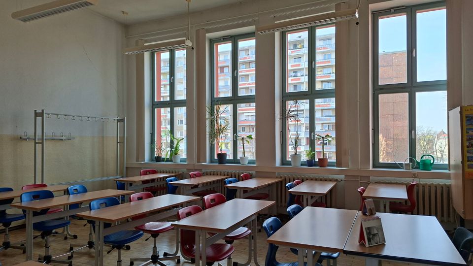 Heller Klassenraum mit mehreren Reihen von Tischen und rot-blauen Stühlen auf Parkettboden, dazu Deckenleuchten und eine Garderobenleiste an der Wand. Große, hohe Fensterfront mit Fensterbänken, auf denen Topfpflanzen und Gießkannen stehen; draußen sind Wohnblocks zu sehen.