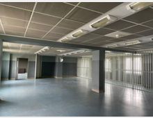 Großer, leerer Innenraum mit grauen Wänden und blau-grauem Boden, getragen von Stützpfeilern und einer abgehängten Decke mit langen Leuchtstofflampen. Rechts befindet sich ein Bereich mit Metallgitter-Trennwänden vor Fensterfronten, links sind mehrere offene Türdurchgänge zu weiteren Räumen.
