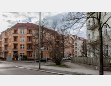 Straßenecke in einem Wohnviertel mit mehrgeschossigen Altbauhäusern in warmen Farben (Orange und Rosa) und dunkelroten Dächern. Im Vordergrund ein kleiner Gehweg mit Bäumen, Verkehrszeichen und parkenden Autos.