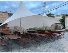 Offener Außenbereich mit mehreren Holzbänken und Tischen auf einem gepflasterten Boden. Ein großes, weißes Sonnensegel überspannt den Sitzbereich, im Hintergrund sind eine Backsteinmauer und weitere Gebäude sowie ein Mobilfunkmast sichtbar.