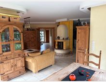 Wohnzimmer in rustikalem Stil mit Holzmöbeln, darunter ein großer Schrank mit Glastüren, ein Esstisch und eine Kommode. Im Hintergrund ein offener Kamin mit runder Einfassung, gelb gestrichene Wände und Fliesenboden.