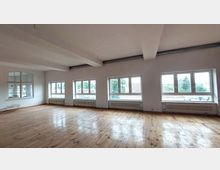 Leerer Raum mit großen Fenstern an zwei Wänden, die viel Tageslicht hereinlassen. Der Boden ist aus hellem Holz, die Wände sind weiß, und Heizkörper befinden sich unter den Fenstern.