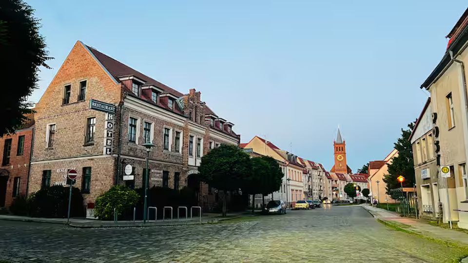 Kopfsteingepflasterte Straße in einer Kleinstadt mit historischen Backstein- und Putzhäusern, darunter ein Eckgebäude mit Hotelbeschilderung. Im Hintergrund ist ein roter Kirchturm mit Spitze am Ende der Straße zu sehen.