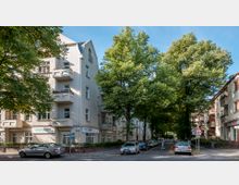 Mehrfamilienhäuser an einer ruhigen Straßenkreuzung mit großen Bäumen, die den Gehweg und die Straße teilweise beschatten. Im Erdgeschoss eines Gebäudes befinden sich Geschäftsräume mit Schildern, während auf der Straße parkende Autos sichtbar sind.