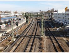 Bahnhofsbereich mit mehreren Gleisen, flankiert von einem überdachten Bahnsteig auf der linken Seite und einem weißen Gebäude auf der rechten Seite. Oberleitungen und metallene Stützpfeiler sind sichtbar, im Hintergrund eine Stadtlandschaft mit Kränen und Gebäuden.