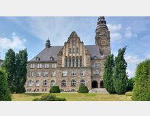 Historisches Gebäude mit einer Fassade aus Naturstein, zahlreichen Fenstern und einem großen Ziergiebel in der Mitte; rechts erhebt sich ein Turm mit Uhr und spitzem Dach. Im Vordergrund befinden sich gepflegte Grünflächen und symmetrisch angeordnete Bäume.