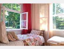 Ein gemütliches Zimmer mit einem kleinen Bett, das mit einer bunten Tagesdecke bedeckt ist, und einem geöffneten Fenster, durch das grüne Bäume und ein sonniger Garten sichtbar sind. Der Raum ist mit roten und beigen Vorhängen sowie einem Rattanhocker dekoriert.