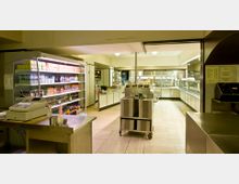 Selbstbedienungskantine mit moderner Ausstattung: Im Vordergrund ein Verkaufstresen mit Kasse und Kühlschrank voller Getränke und Snacks, im Hintergrund eine Theke mit Warmhaltebehältern und ein Blick in die Kücheneinrichtung. Der Raum ist gut beleuchtet und bietet eine klare Aufteilung der Bereiche.
