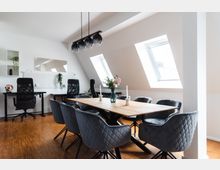 Heller Besprechungsraum unter dem Dach mit schrägen Decken und zwei Dachfenstern, einem Holztisch mit grauen Polsterstühlen sowie modernen Hängelampen. Im Hintergrund befinden sich zwei Schreibtische mit schwarzen Bürostühlen und dekorativen Pflanzen.