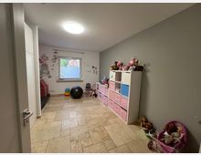Ein Kinderzimmer mit hellen Wänden und einem beigefarbenen Fliesenboden. Links ein Sitzbereich mit einem Wandtattoo, rechts ein Regal mit rosa und blauen Aufbewahrungsboxen sowie Spielzeug, darunter Puppen und ein Kinderwagen.