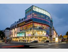 Moderne Einkaufszentrum-Fassade bei Abenddämmerung, mit Glasfronten und neonbeleuchteten Ladenbeschriftungen wie „Primark“ und „Schloss-Straßen-Center“. Im Vordergrund eine belebte Straßenkreuzung mit Autos, Fußgängern und Straßenschildern.
