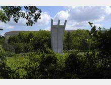 Eine Betonskulptur mit drei nach oben geneigten Säulen, bekannt als Luftbrückendenkmal, steht inmitten einer grünen Parklandschaft mit Büschen und Bäumen; im Hintergrund sind Teile eines Gebäudes und ein leicht bewölkter Himmel zu sehen.