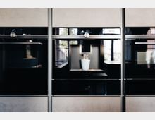 In der Mitte einer Reihe moderner Einbaugeräte befindet sich eine integrierte Kaffeemaschine mit einer weißen Tasse, die gerade befüllt wird. Die Gerätefronten sind schwarz mit Edelstahlakzenten, und im Hintergrund sind verschwommene Fenster und Tageslicht sichtbar.