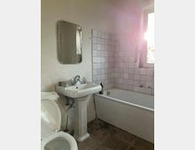 Ein einfaches Badezimmer mit sandfarbenen Wänden, einem Waschbecken mit Wandspiegel, einer weißen Badewanne mit gefliester Verkleidung und einem offenen Fenster, durch das Sonnenlicht hereinfällt. Im Vordergrund ist eine weiße Toilette auf einem Steinboden zu sehen.