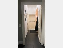 Schmaler Flur mit dunklem Fliesenboden und weißen Wänden, der zu einer beigen Tür mit einem Sicherheitsbügel führt. An der rechten Wand hängt ein schwarzer Mantel, links sind ein Wandspiegel und Dekoration zu sehen.