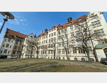 Großer Innenhof vor einem mehrstöckigen historischen Gebäude mit heller Fassade, vielen rechteckigen Fenstern und roten Ziegeldächern mit kleinen Dachaufbauten. Im Vordergrund stehen kahle Bäume und eine gepflasterte Rasenfläche, links ist ein Flügel mit Fachwerkdetails zu sehen.