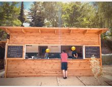 Ein hölzerner Imbissstand mit offenem Verkaufsfenster, hinter dem Mitarbeiter arbeiten. Links und rechts des Fensters befinden sich schwarze Tafeln mit einer handgeschriebenen Pizzaspeisekarte. Im Hintergrund stehen Bäume und es herrscht sonniges Wetter.