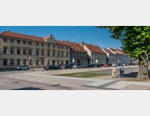 Straßenszene mit einer Reihe von historischen Wohnhäusern mit roten Ziegeldächern, davor geparkte Autos auf einer gepflasterten Straße. Im Vordergrund ein Weg mit Grünflächen und zwei Personen, die spazieren gehen, rechts ein großer Baum.