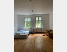 Ein schlicht eingerichtetes Zimmer mit Holzboden und weiß gestrichenen Wänden. Es gibt zwei große Fenster mit Blick auf Bäume, ein bodennahes Bett, einen beistelltisch mit Lampe und zwei gelbe Sessel am rechten Rand.