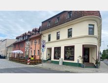 Mehrstöckige Häuserreihe mit roten Ziegeldächern und farbenfrohen Fassaden in einer urbanen Straße, darunter ein cremefarbenes Gebäude mit Schaufenstern eines Ladens. Im Vordergrund ein Gehweg mit Pflanzkübeln und Straßenschilder.