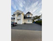 Zweistöckiges Wohnhaus mit hellem Putz, schwarzem Dach und symmetrischem Design, inklusive Säulen und Balkon über dem Eingangsbereich. Das Haus steht auf einem gepflasterten Hof mit einem geparkten Auto und ist von Bäumen im Hintergrund umgeben.