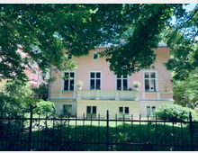 Rosa Haus im klassischen Stil mit großen weißen Fenstern, umgeben von einem gepflegten Garten mit Bäumen im Vordergrund und einem schmiedeeisernen Zaun. Im Hintergrund sind weitere Gebäude und dichtes Grün zu sehen.