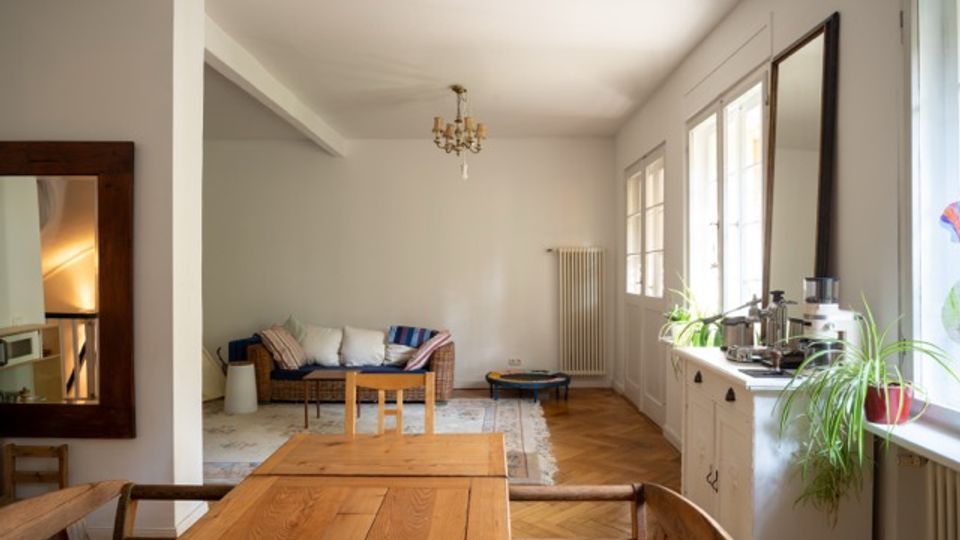 Heller Wohn- und Essbereich mit Holztisch im Vordergrund, Sitzbank und Teppich im Hintergrund sowie Parkettboden und Kronleuchter an der Decke. Rechts sorgen große Fenster und eine verglaste Tür für viel Tageslicht; darunter steht ein Sideboard mit Pflanzen und Dekoration, daneben ein hoher Heizkörper.