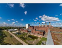 Industriekomplex aus roten Backsteingebäuden mit hohen Schornsteinen und einem weißen Turm, umgeben von einer grünen Parkanlage mit Teichen und einem Gewächshaus; der Himmel ist blau mit einzelnen Wolken.