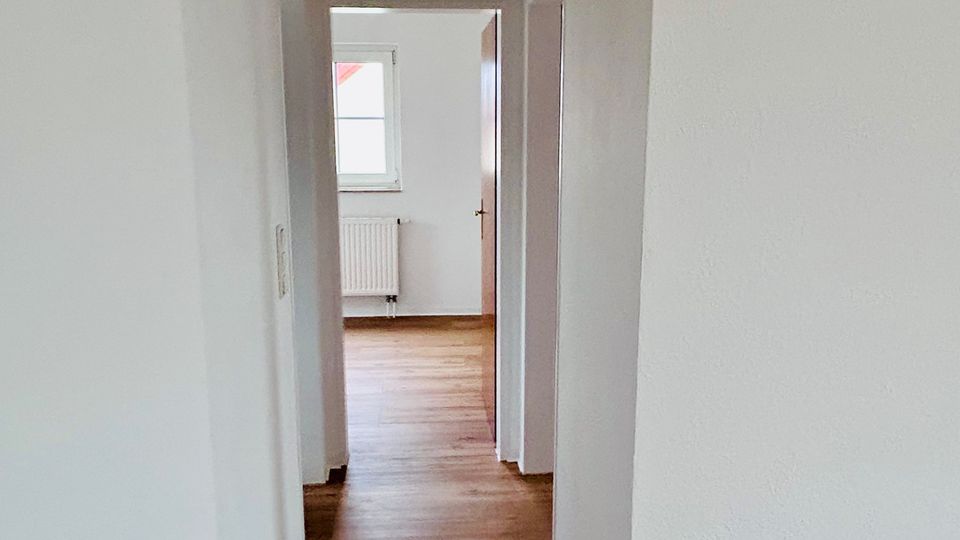 Schmaler, heller Wohnungsflur mit weiß verputzten Wänden, Holzboden und einem Rundbogen-Durchgang. Am Ende führt eine offene Tür in einen Raum mit Fenster und darunterliegendem Heizkörper.