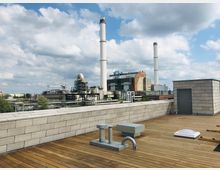 Dachterrasse mit Holzplanken und technischen Installationen, im Hintergrund eine Industrieanlage mit großen Schornsteinen und Gebäuden verschiedener Bauweisen unter einem teils bewölkten Himmel.