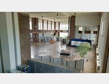 Großer Innenraum einer ehemaligen Industriehalle mit hohen Decken, großen Fenstern und Backsteinwänden. Der Raum ist mit Schautafeln, einigen technischen Geräten und Modellen bestückt; ein gefliester Boden und mehrere Pflanzen sind sichtbar.