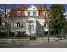 Zweistöckige Stadtvilla mit heller Fassade, roten Dachziegeln und weißen Klappläden vor den Fenstern, umgeben von einem kunstvollen schwarzen Metallzaun. Vor dem Gebäude befinden sich Bäume und ein Gehweg entlang der Straße.