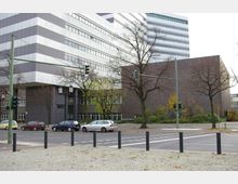 Straßenkreuzung mit Fußgängerüberwegen vor einem modernen Bürogebäude mit dunklen und weißen horizontalen Fassadenstreifen sowie einem angrenzenden Backsteinbau; kahle Bäume und parkende Autos im Vordergrund.