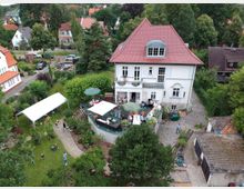 Großes, zweistöckiges Wohnhaus mit rotem Satteldach in einem grünen Gartenumfeld. Auf der Terrasse und im Garten befinden sich Pavillons, Sitzbereiche und mehrere Personen; umgeben von Bäumen, Rasenflächen und gepflasterten Wegen.
