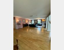 Wohnzimmer mit einem hellen Holzfußboden, großen Fenstern mit Blick ins Grüne und modernen Möbeln, darunter ein dunkelgrünes Sofa und mehrere Lampen. Wände sind teils in Altrosa gestrichen, mit dekorativen Bildern und einem Sideboard an der Wand.