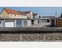 Ein offener Garagenhof mit mehreren nebeneinander angeordneten Garagentoren in unterschiedlichen Farben. Im Hintergrund sind mehrstöckige Wohnhäuser mit roten und gelben Dächern sowie ein einziger Turm sichtbar.