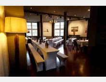 Innenraum eines Restaurants mit langen, weiß gedeckten Tafeln und Bänken sowie weiteren Tischen mit Stühlen auf dunklem Holzboden. Große Sprossenfenster, schwarze Stützsäulen, eine Kronleuchte und Pendelleuchten prägen den Raum.