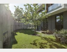 Grüne Gartenfläche mit einem jungen Baum in der Mitte, umgeben von einer hohen grauen Mauer im Hintergrund. Rechts im Bild ein modernes Wohngebäude mit großen Glasfenstern und Balkonen.