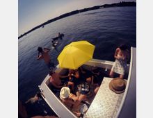 Menschen entspannen auf einem kleinen Boot auf einem ruhigen See, teils sitzend unter einem gelben Sonnenschirm, während andere im Wasser schwimmen. Im Hintergrund sind Bäume und ein flacher Horizont zu sehen.