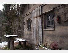 Holzhütte mit rustikalen Holztüren und kleinen Fenstern, umgeben von winterlicher Waldlandschaft. Im Vordergrund steht ein einfacher Holztisch mit Bank, teilweise mit Schnee bedeckt, daneben eine Pflanzenschale mit Heidekraut.