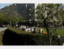 Eine städtische Parkszene mit einer von Hecken eingefassten Grünfläche, Blumenbeeten und einer Sitzbankreihe, auf der Menschen in der Sonne sitzen. Im Hintergrund sind mehrstöckige Wohngebäude mit Geschäften im Erdgeschoss zu sehen.