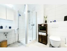 Modernes Badezimmer mit weißen Fliesenwänden und grauem Fliesenboden. Es verfügt über eine Glasdusche, ein Waschbecken mit Spiegel, eine wandmontierte Toilette und einen Holzschrank mit Handtüchern und Dekoration.