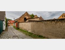 Schmale, gepflasterte Gasse in einem Dorf, umgeben von kleinen Häusern mit Ziegeldächern, darunter auch ein Backsteinbau. Eine verputzte Steinmauer dominiert die rechte Seite, und im Hintergrund ist ein orangefarbenes Wohnhaus zu sehen.