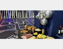 Buffet-Tisch mit Käse-, Wurst- und Obstplatten sowie Snacks vor einer dunkelblauen Wand mit silberner Ballondekoration; im Hintergrund ein Indoor-Spielbereich mit markierten Spielfeldern und kleinen Roboterfahrzeugen auf dem Boden.