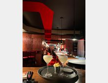 Eine stilvolle Bar mit gedämpftem Licht und rotem Dekor, darunter eine auffällige rote Deckenstruktur. Im Vordergrund stehen zwei Cocktails auf einem Tablett, daneben eine kleine Schale mit Popcorn, während Gäste im Hintergrund an einem langen Tresen sitzen.