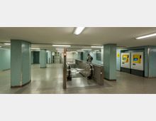Unterirdischer U-Bahn-Bereich mit blauen gefliesten Wänden, hellgrauem Boden und einer Rolltreppe in der Mitte. Auf der rechten Seite befinden sich zwei gelbe Fahrkartenautomaten, im Hintergrund ist ein Fahrstuhl zu sehen.