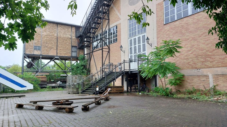 Backstein-Gebäude eines ehemaligen Industriekomplexes mit großen Fenstern, einer Metalltreppe und umliegenden Pflanzen. Im Vordergrund gepflasterter Hof mit alten Holzgestellen und Metallkonstruktionen.